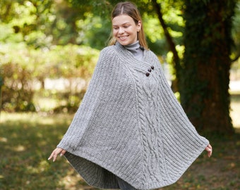 May include: A gray knitted poncho with a cable knit pattern and two buttons. The poncho is worn by a person standing in a park.