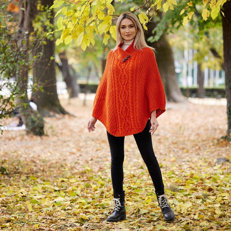 May include: A woman wearing a bright orange knitted poncho with a cable knit pattern and black leggings. She is standing in a park with fallen leaves on the ground.