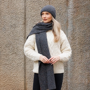 May include: A woman wearing a gray knit beanie, scarf, and cream-colored cable-knit sweater. The scarf and beanie are a speckled gray. The sweater has a classic cable-knit design. The outfit is suitable for cold weather.