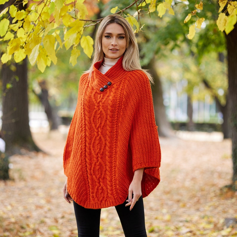 May include: A bright orange knitted poncho with a cable knit pattern and two brown buttons. The poncho is worn by a person standing in a park with fall foliage in the background.