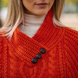 May include: A close-up of a bright orange knitted sweater with a cable knit pattern and three brown buttons on the collar.
