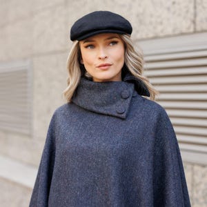 Peut inclure: Une femme portant un poncho en laine bleu foncé avec un col montant et deux boutons. Elle porte une casquette plate noire.