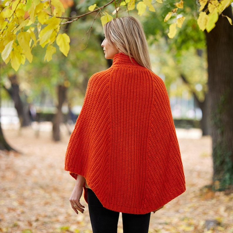 May include: A woman wearing a bright orange knitted poncho in a park setting. The poncho has a turtleneck and a loose, flowing fit.