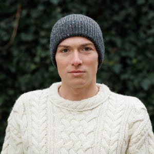 May include: A gray knit beanie with flecks of color. The ribbed hat is worn by a person. The person is wearing a cream-colored cable-knit sweater. The background is a blurred green.