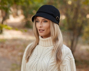 May include: A black cloche hat with a small brim and a decorative gold-colored buckle. The hat is made of a textured fabric. The person is wearing a cream-colored cable-knit turtleneck sweater.