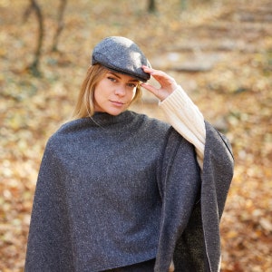 May include: A woman wearing a grey tweed poncho with a grey tweed newsboy cap. The poncho has a high neckline and a loose fit. The woman is standing in a woodland setting.