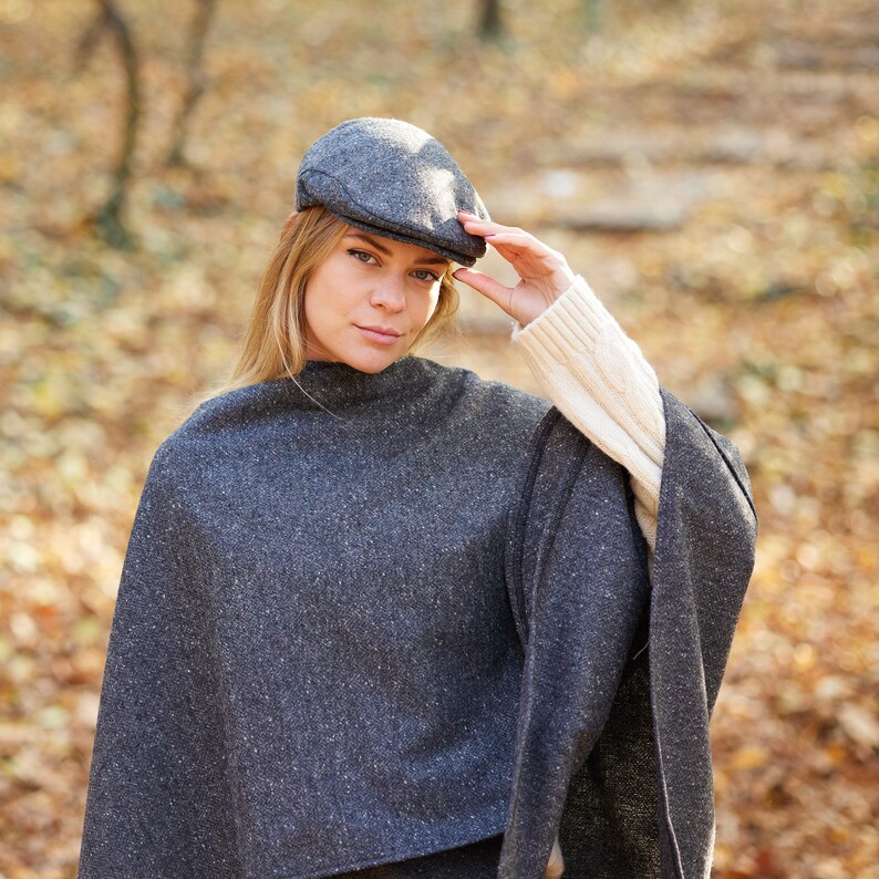 May include: A woman wearing a grey tweed poncho with a grey tweed newsboy cap. The poncho has a high neckline and a loose fit. The woman is standing in a woodland setting.