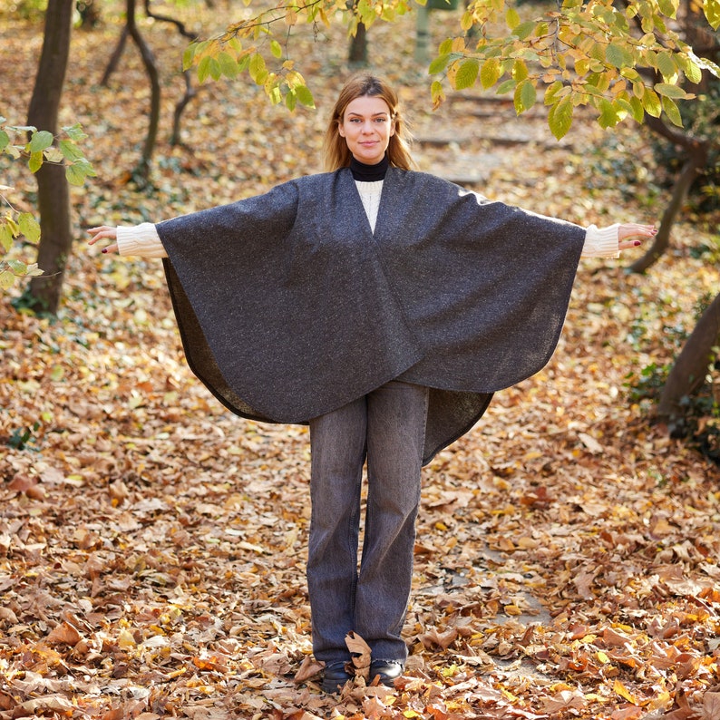 May include: A woman wearing a grey wool cape with a white sweater underneath. She is standing in a woodland with fallen leaves on the ground.