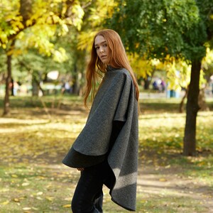 May include: A woman wearing a grey wool poncho in a park setting. The poncho has a long, flowing design and is made of a soft, warm material. The woman is standing in a park with trees and fallen leaves in the background.