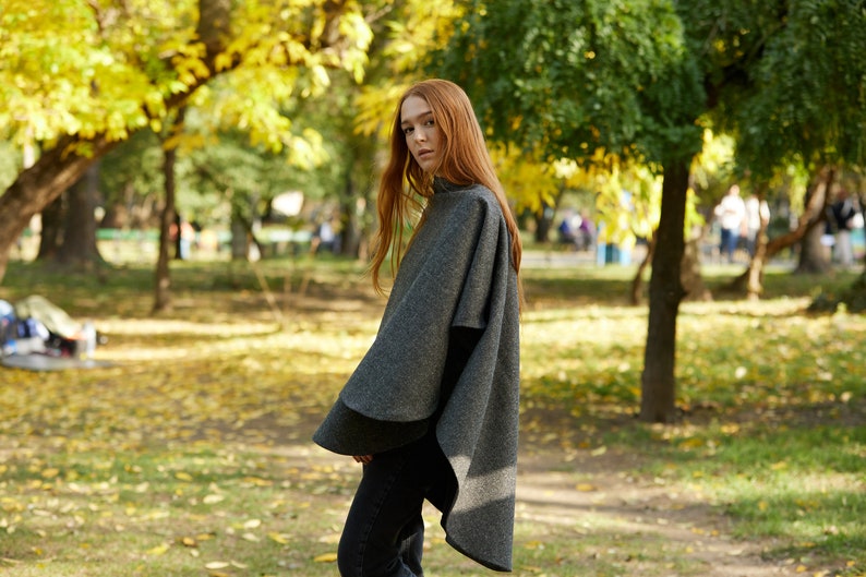 May include: A woman wearing a grey wool poncho in a park setting. The poncho has a long, flowing design and is made of a soft, warm material. The woman is standing in a park with trees and fallen leaves in the background.