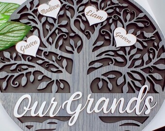 May include: A gray wooden family tree with the words "Our Grands" at the bottom. There are three white hearts with names on them: Madison, Liam, and Gavin.