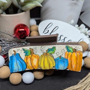 May include: A brown hair clip featuring a hand-painted design of pumpkins in shades of blue, orange, and yellow. The clip has a rectangular shape with a decorative pumpkin theme, perfect for fall.