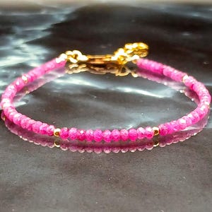 May include: A delicate bracelet featuring small, faceted hot pink beads strung together with tiny gold-toned metal spacers. The bracelet is finished with a gold-toned metal lobster claw clasp and a short extender chain at the back for adjustment.