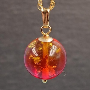 May include: A gold-colored chain necklace with a pendant. The pendant is a transparent red sphere with gold-colored flecks inside. The necklace hangs against a dark gray background.
