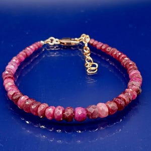 May include: A bracelet composed of small, faceted ruby beads in shades of pink and red. The bracelet is strung on a gold-coloured chain with a lobster clasp. The background is a solid, deep blue.