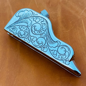 May include: A silver-colored metal tool with an intricate floral design engraved on the surface. The tool has a rounded, curved shape and a small knob on the top.