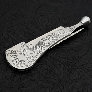 May include: A silver pipe tool with a decorative floral design. The tool has a small, round end and a long, thin blade.