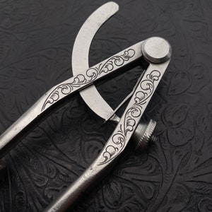 May include: A silver compass with intricate floral engravings. The compass has a hinged arm and a knob for adjusting the width of the compass.