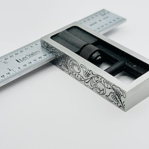 May include: A silver metal try square with a black adjustment knob and a decorative floral pattern engraved on the surface. The ruler is marked with inches and the text "Lee Valley" is printed on the ruler.