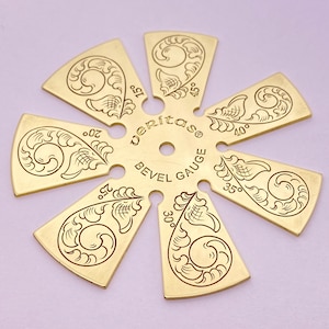 May include: A gold-colored Veritas bevel gauge with a central hole and six radiating arms. Each arm is engraved with a floral design and a degree marking, ranging from 15 to 45 degrees. The text "Veritas BEVEL GAUGE" is etched in the center.