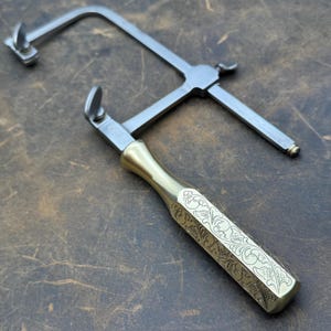 May include: A jeweler's saw with a brass handle and a silver-colored frame. The handle is engraved with a floral pattern. The saw is designed for precision cutting in jewelry making and other crafts.