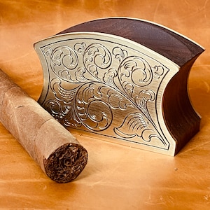 May include: A decorative wooden and metal cigar holder with an intricate floral design. The holder is made of dark wood and has a silver-colored metal top with a floral pattern.