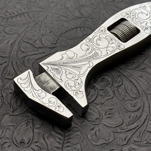 May include: A silver adjustable wrench with an intricate floral design engraved on the handle and jaw. The wrench is lying on a black textured surface.