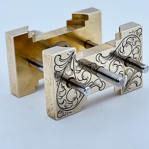 May include: A brass tool with intricate floral engravings and two silver screws. The tool has a rectangular shape and is designed for holding and securing objects.