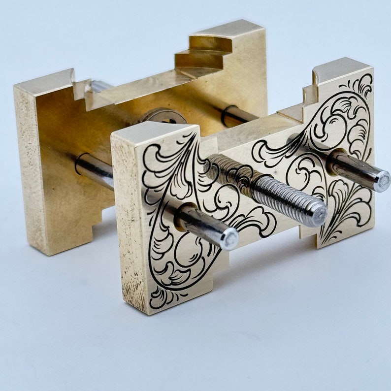 May include: A brass tool with intricate floral engravings and two silver screws. The tool has a rectangular shape and is designed for holding and securing objects.