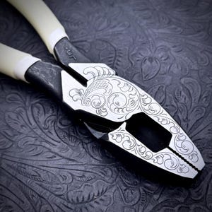 May include: Close-up of a pair of pliers with a decorative floral engraving on the metal surface. The pliers have black jaws, a silver-colored center, and off-white handles. The background is a dark, textured surface.
