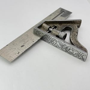 May include: A vintage combination square with a silver finish. The square features an etched floral design along the edges and a ruler with inch measurements. The ruler is inserted into the square's body. The tool is designed for woodworking and metalworking.