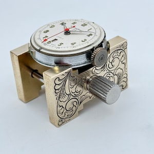 May include: A vintage watch with a white face and black markings is held in a brass watchmaker's tool with an engraved floral design.