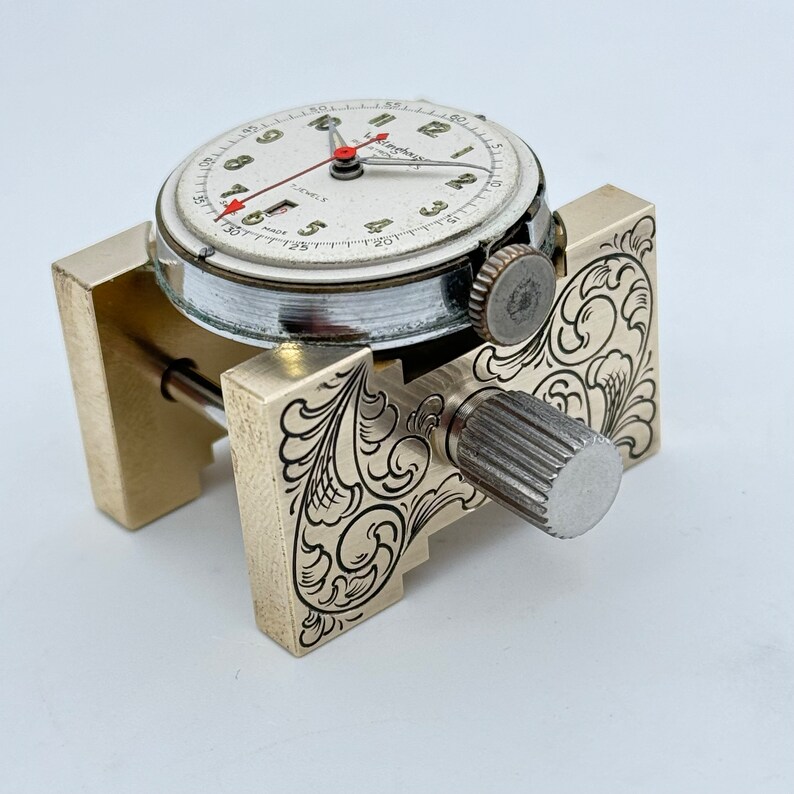 May include: A vintage watch with a white face and black markings is held in a brass watchmaker's tool with an engraved floral design.