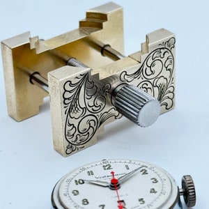 May include: A silver watchmaker's clamp with an engraved floral design. The clamp has a knob with a knurled surface. The clamp is used to hold a watch face in place while working on it.