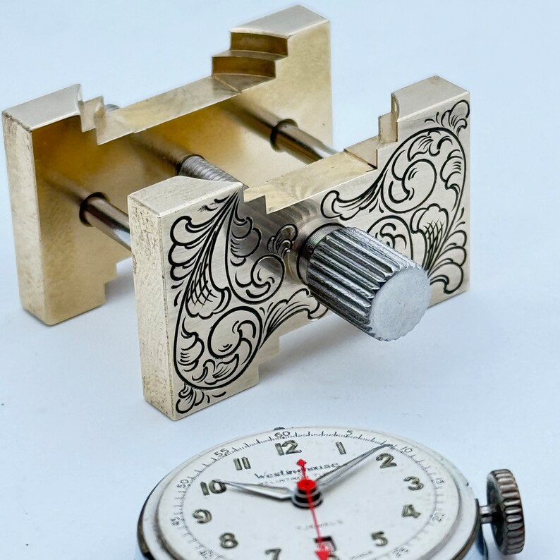 May include: A silver watchmaker's clamp with an engraved floral design. The clamp has a knob with a knurled surface. The clamp is used to hold a watch face in place while working on it.