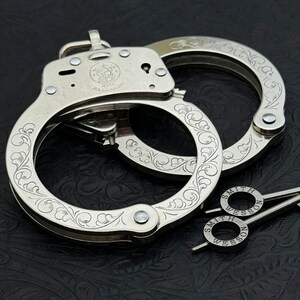 May include: Silver-colored handcuffs with intricate floral engraving. The handcuffs are marked "SMITH & WESSON" and include two keys. The design features detailed patterns on the cuffs and the key rings.