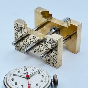 May include: A brass watchmaker's vise with intricate floral engravings. The vise has two adjustable screws and a lever for clamping. A vintage watch face with black hands and white numerals is visible in the foreground.