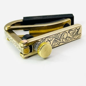 May include: A brass guitar capo with intricate floral engraving. The capo features a black rubber pad and a gold-colored adjustment knob. The design includes a spring mechanism for secure placement on the guitar neck. The capo is designed to change the pitch of the guitar.