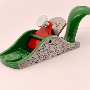 May include: A green wood plane with a red adjustment knob and a silver blade. The plane has a decorative engraved design on the side. The blade is labeled "Kunz".