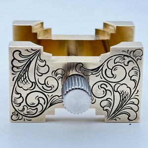 May include: A gold-colored metal tool with a decorative engraved design. The tool has a knob on the front and a square opening in the center.