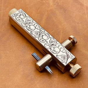 May include: A brass tool with an engraved floral design. The tool has a rectangular body with a screw at each end and a small blade extending from the side. The tool is set against a brown leather background.