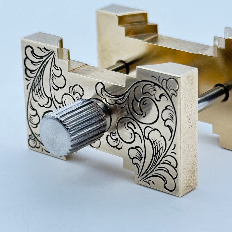 May include: A brass watchmaker's clamp with an intricate engraved floral design. The clamp has a silver knob for adjusting the pressure.