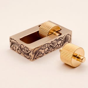 May include: A rectangular brass block with an open center and intricate black scrollwork engravings along the side. The block features a circular, knurled brass knob set within the opening, accompanied by a separate, matching knurled brass knob.