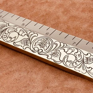 May include: Close-up of a metal ruler with engraved floral designs. The ruler has inch markings from 4 to 7. The engraved design is dark against the lighter metal. The ruler is on a brown surface.