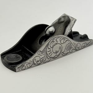 May include: A black metal hand plane with a decorative engraved floral design. The plane has a metal blade and a wooden handle.