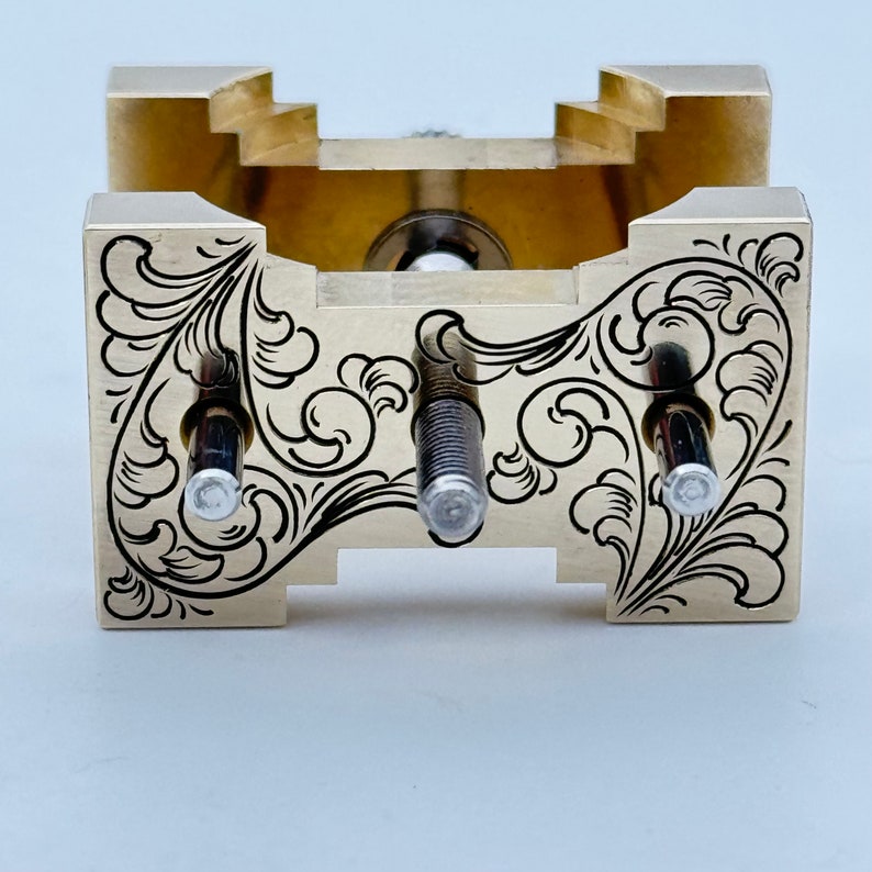 May include: A gold-colored metal tool with intricate engraved designs. The tool has three adjustable screws and a square base.