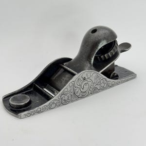 May include: A small, antique metal hand plane with a dark gray finish. The plane features ornate, engraved floral patterns along the sides. The blade is visible within the body of the plane, and a small knob is on the front.