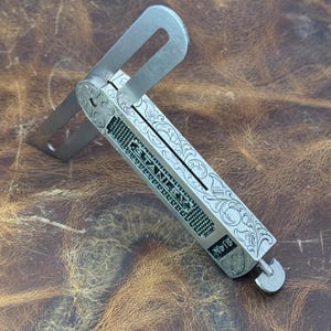 May include: A vintage Stanley No. 18 combination square with ornate engraved detailing. The tool is silver-toned with a rectangular body and an adjustable blade. The body features the "STANLEY" brand name.