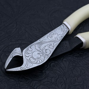 May include: A pair of pliers with a white handle and a decorative floral pattern etched into the metal. The pliers are lying on a black textured surface.
