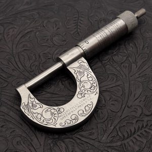 May include: A silver-toned metal micrometer featuring intricate scrollwork engravings on the frame. The tool includes a knurled adjustment knob and a barrel with printed decimal equivalents. Text on the frame reads: MILLERS FALLS CO NO 112 GREENFIELD, MASS. U.S.A.